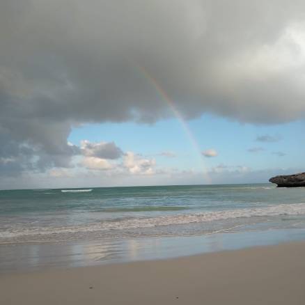 A wave breaking on a beach beneath a blue sky, thundercloud, and rainbow