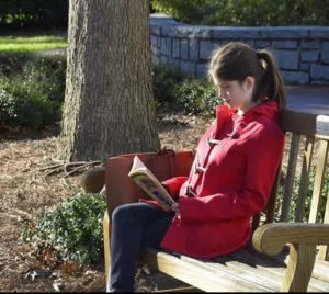 Deanna, a brown-haired girl in a red peacoat, sits on a park bench reading Frankenstein. 