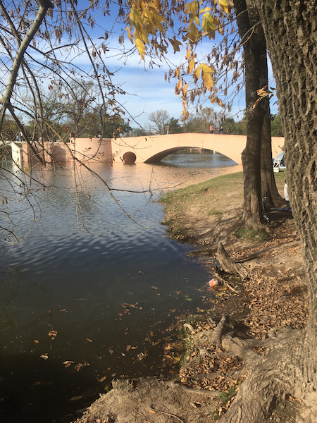 A low, flat pink bridge over a stream. The riverbank and trees are to the right.