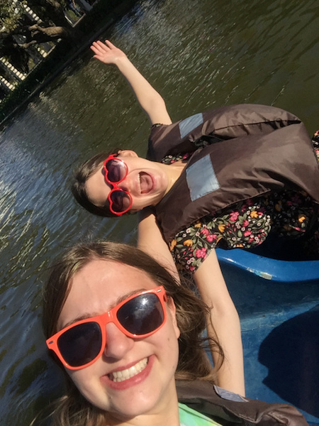 Deanna and a friend wear sunglasses and life vests while riding a paddleboat. Their arms are splayed in celebration.