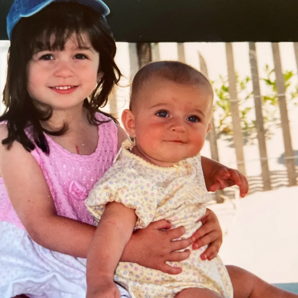 To the left, a brown-haired little Deanna wraps her arms around her baby sister. They’re sitting on a deck outside. It’s a few weeks after Deanna’s open heart surgery. This experience inspired Deanna to write Chrysalis.
