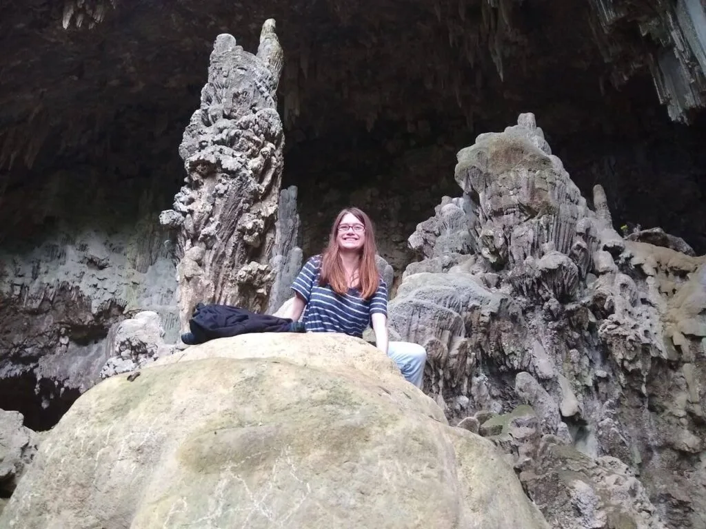 A brown-haired girl sitting atop a boulder in a cave. Behind her are two elaborate stalagmites.