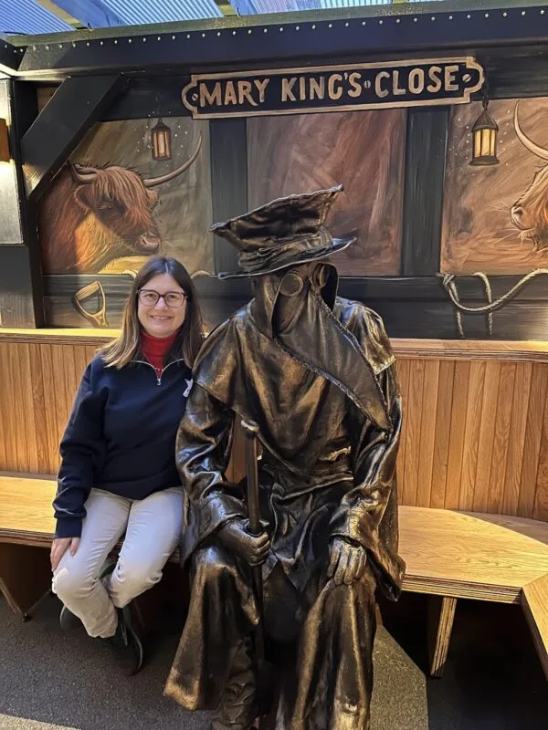 A brown-haired White girl sits on a bench next to a statue of a plague doctor wearing a cloak and long, beak-like mask